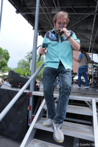Male singer on an outdoor stage, holding a microphone and stepping down the metal stairs, wearing a turquoise shirt with dark floral details beside him on stage.