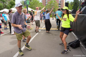Crowd of people dancing and cheering on a city street at an outdoor festival, with a stage on the right and a bright neon-shirted dancer in the foreground.