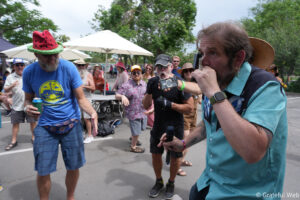 Crowd at an outdoor festival with a man in a teal shirt speaking into a microphone near the camera and others cheering in the background.