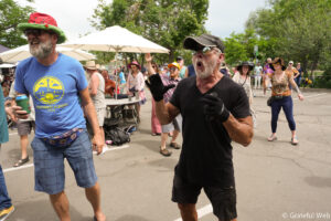 Bearded man in a blue t-shirt and colorful hat dancing at an outdoor festival with a cheering crowd in the background under umbrellas and trees.