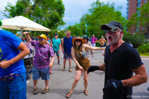 Group of people dancing and having fun at an outdoor street festival on a sunny day, with trees in the background.