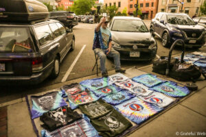Man sits on a folding chair on a city street, displaying colorful graphic T-shirts laid out on the sidewalk for sale.