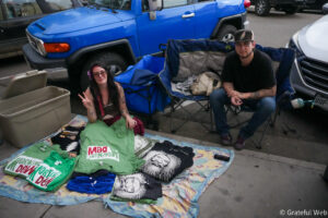 Two young adults sit on a colorful blanket and folding chairs on a sidewalk, surrounded by clothes and bags, with a bright blue Jeep parked behind them.