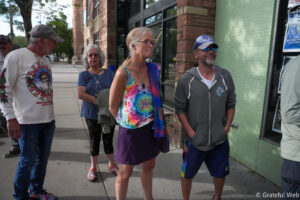 Group of four adults standing on a sidewalk outside a brick storefront, chatting and looking to the left.