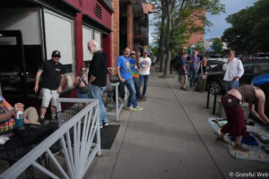 Group of casually dressed people standing along a sidewalk outside a brick storefront, chatting and drinking.