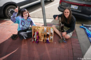 Two friends sit on a brick sidewalk beside a display board of feather earrings and jewelry, one giving a thumbs-up while a guitar rests nearby.