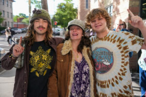 Three friends pose together on a sunny city street, smiling at the camera with arms around each other.