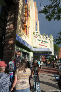 Colorful storefront with a bright marquee, a brick corner, and a crowded sidewalk on a sunny day.