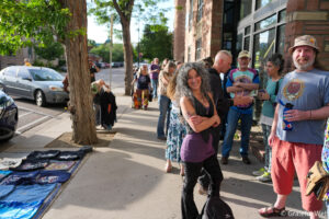 Smiling woman with curly hair and crossed arms standing in a queue outside a brick building, others lined up behind her on a sunny street.