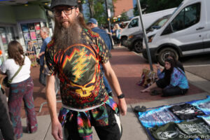 Bearded man in a colorful, psychedelic shirt standing on a city sidewalk.