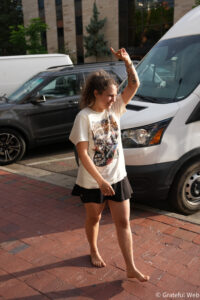 Young girl walking barefoot on a brick sidewalk, raising her right arm with a floral tattoo visible, with vans and a building behind her.