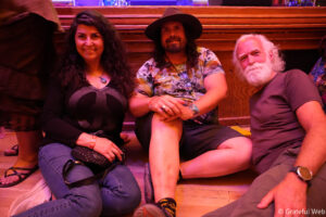 Three friends sit on a wooden stage floor under pink lighting, smiling at the camera.