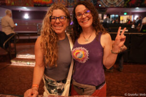 Two smiling women posing for a photo at an indoor event; one with glasses and long curly blonde hair, the other flashing a peace sign.