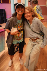 Two women posing for a photo, smiling with their arms around each other in a colorful room with wood floor.