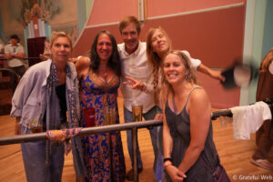 Group of five adults smiling and posing for a photo at a social event, leaning on a railing with drinks in hand.
