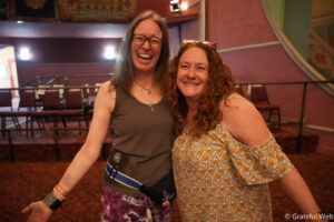 Two smiling women pose closely for a photo in a dim, colorful venue with rows of chairs in the background.
