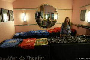 Vendor booth with pink walls selling blue and red t-shirts and pin trays, large round mirror reflecting the room, framed photos on the wall, and a staff member nearby.