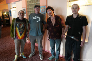 Four men stand side by side in a pink-walled hallway, posing for the photo in casual shirts and jeans.