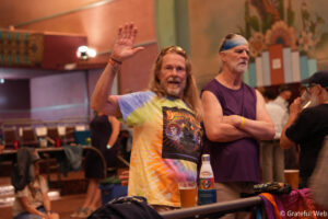 Man with a tie-dye shirt waves to the camera at a crowded indoor event, drinks on the table nearby.