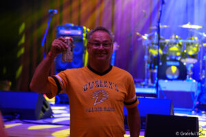 Smiling man in an orange shirt raises a beer can on a colorful concert stage in the background.
