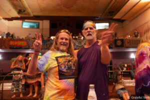 Two men wearing tie-dye shirts pose for the camera in a crowded indoor event, flashing peace signs.