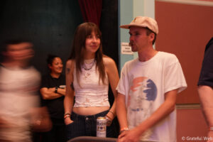 Two young adults stand together at a casual indoor event; woman in a white lace top holds a can, man in a white T-shirt and beige cap talks beside her.