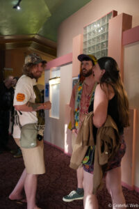 Three people chatting in a pastel hallway at an indoor event: man in a camo cap and beige shorts talks to a bearded man with a colorful scarf and a woman with a brown jacket.