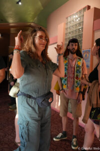 Woman in a green utility jumpsuit gestures with raised arms while chatting with others at an indoor event. Korean-inspired colorful vest man stands nearby.