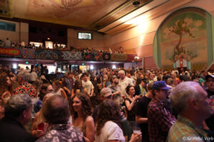 Crowded indoor event in a large hall with colorful banners along the balcony railing and a mural on the wall.