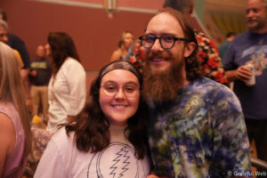 Two friends pose together in a crowded indoor party, smiling at the camera (woman with glasses in front, man with beard beside her).