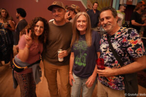 Four friends pose for a close-up group photo at a lively indoor party, holding drinks and smiling at the camera.