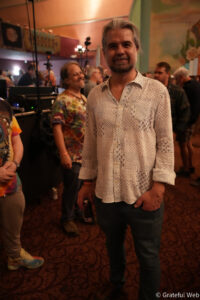 Smiling man with shoulder‑length gray hair and a patterned white shirt standing in a crowded indoor event space with warm lighting in the background.