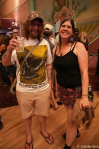 Two smiling adults posing at a crowded indoor party, with colorful mural in the background and wooden floor.