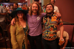 Three friends pose and smile at a crowded indoor party; woman on the left wears a teal hat and glasses, center man in a purple shirt, right man in a tie-dye shirt with headphones around his neck.