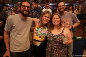 Four friends posing for a photo at a crowded party; two women in front, drinks in hand, smiling at the camera.