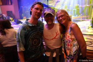 Three friends posing for a photo at a music venue with vibrant stage lights behind them, smiling.