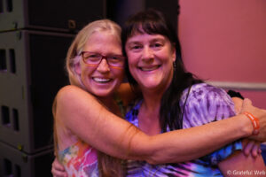 Two women hug and smile at the camera in a dimly lit room, one wearing red glasses and tie-dye clothing.