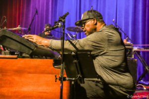 Pianist in a gray hoodie plays a keyboard on a stage with a purple curtain backdrop, microphone in front, drummer in the background.