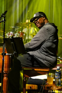 Keyboardist in a gray shirt and cap smiles while playing a keyboard on stage; a drummer and drums appear in the back under green lighting.