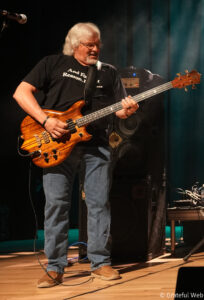 Male bass guitarist on stage, playing a brown striped electric bass with a natural wood finish, wearing a black shirt and jeans under stage lights.