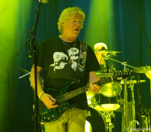 Guitarist on stage under bright yellow-green lighting, playing a dark electric guitar with a patterned strap; drums visible in the background.