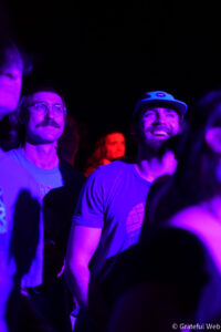 Two men in a dark crowd bathed in purple light; one with glasses and a mustache, the other smiling wearing a cap at a night event