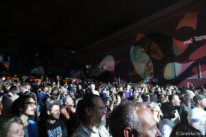 Crowd packed into an indoor concert venue, people facing the stage with colorful mural on the right wall.