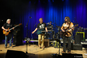 Band performing on a stage with blue backlights; three guitarists in front and a drummer in the back.