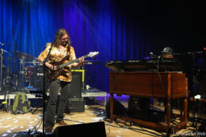 Male guitarist in a tie-dye shirt plays a sunburst electric guitar on a lit stage with a blue curtain backdrop and drums to the left, keyboardist to the right nearby.