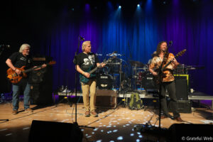 Four musicians on a stage performing live, with two guitarists at the front and a drummer in the back under blue and purple stage lights.