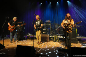Three musicians on a stage performing live: two guitarists at front with one drummer in the back, colorful stage lights.