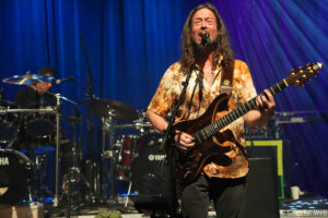 Musician singing into a microphone while playing an electric guitar on stage, with a drummer in the background and blue lighting behind him.