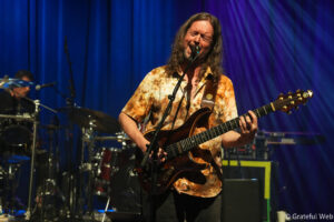 Male guitarist in a tie-dye shirt sings into a microphone while playing an electric guitar on a blue-curtained stage; drummer visible in background.