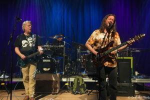 Two musicians performing on a stage: a guitarist with light hair in a black shirt on the left and a long-haired guitarist/vocalist in a tie-dye shirt on the right, drums in the background under blue-purple stage lighting.